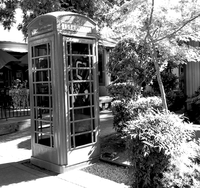 Black-and-white photograph of two vintage phone booths beside a garden path and café.